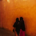 Two women in black walk down a sunlit alley.