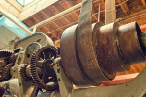 Vintage machine parts in an old factory during daylight hours with wooden beams visible - A view of metal gears and pulleys from an old machine inside a factory. Sunlight enters through a window above, highlighting the machine's design.