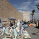 A market displays textiles in front of ancient walls.