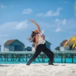 Woman practicing yoga on a sandy beach by the ocean.