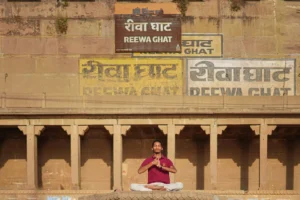 In this serene image, a teenage Indian boy is seen practicing yoga at one of the iconic ghats of Varanasi during sunrise. He sits in a meditative posture with legs crossed in padmasana (lotus position) on a stone platform. His eyes are gently closed, and his hands are joined in a yogic gesture (Anjali Mudra) near his chest, radiating calmness and inner peace. Dressed in a maroon polo shirt and light trousers, the boy is bathed in the soft golden morning light. Behind him, the sunlit architecture of the ghat stands tall, with intricate designs and subtle activity in the background, capturing th
